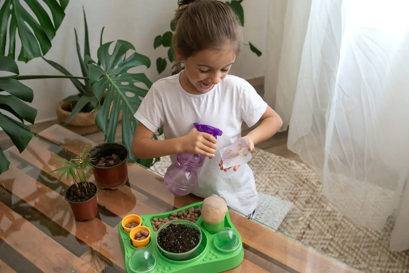 Lustiges kleines Mädchen als Gärtnerin mit Pflanzen im Zimmer zu Hause, gießt und pflegt Zimmerpflanzen, pflanzt Blumen um.
